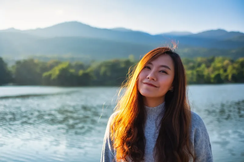 Asian woman standing at golden hour by lakeside, peaceful demeanor