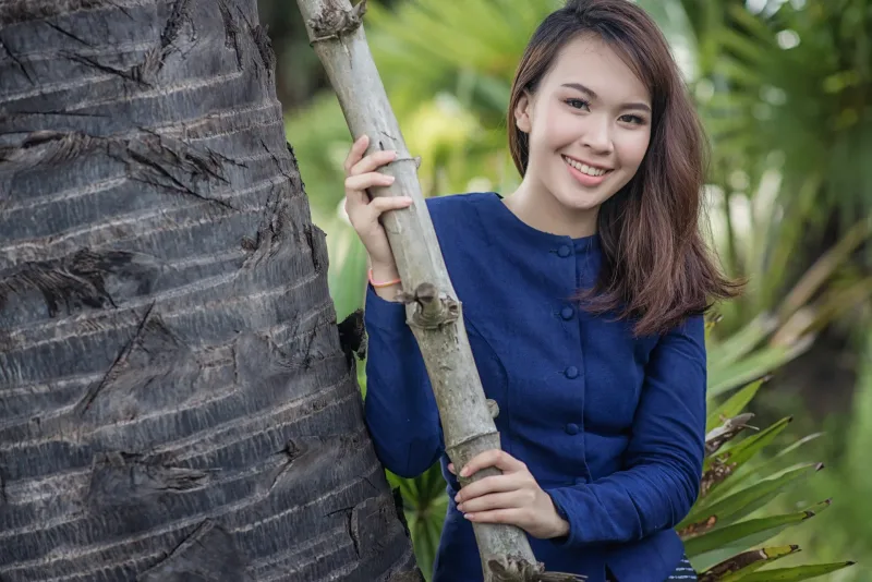 Woman in blue áo dài leaning against tree in tropical setting