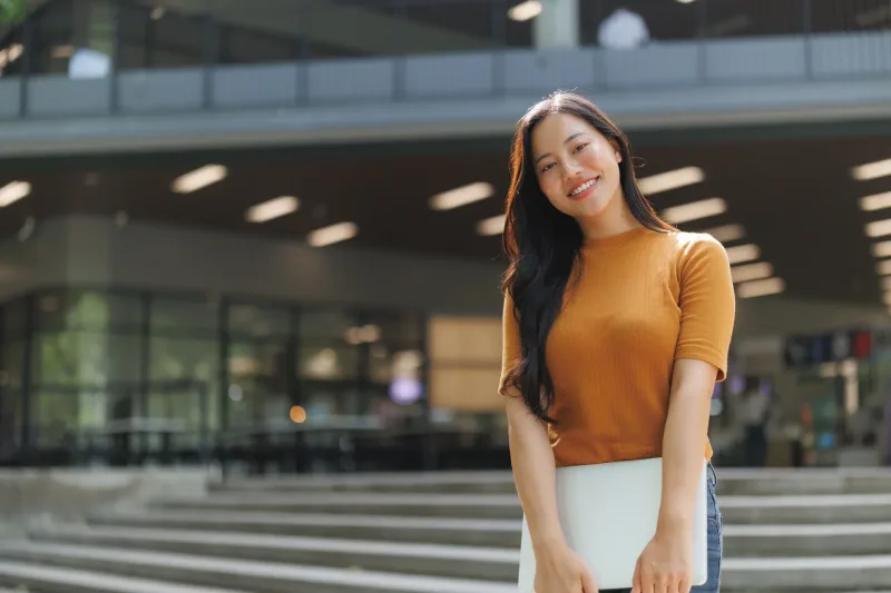 Young woman holding laptop outdoors with casual confidence, symbolizing modern educational systems