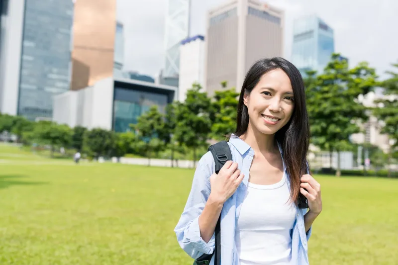 Woman smiling in urban park with city skyline, student with backpack