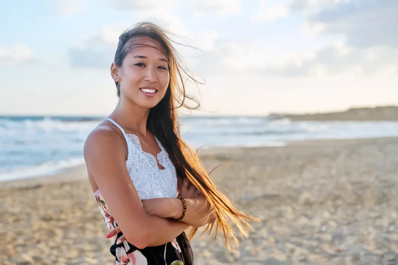 Asian woman smiling outdoors at golden hour on a beach, radiating warmth and genuine care.